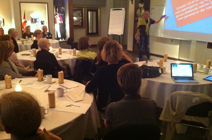 En gruppe mennesker sidder ved borde og ser op mod en underviser En gruppe mennesker sidder ved borde og ser op mod en underviser